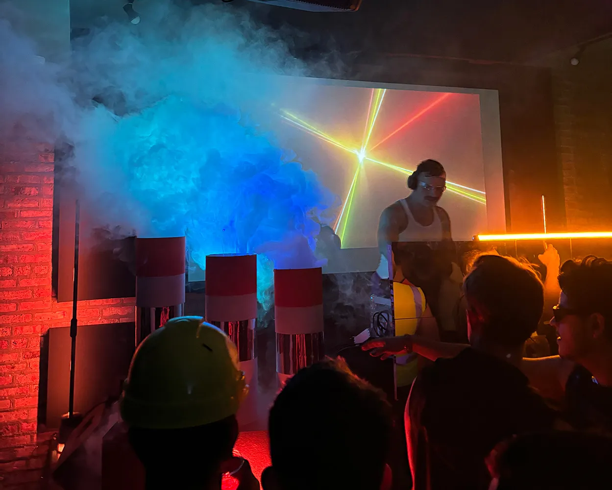 A DJ plays music, cueing a track in his headphones, while there is a set of three smoke stacks that are pumping fog onto the dancefloor